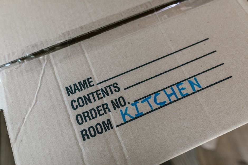 Two women engaged in a home relocation process are standing indoors among several stacked cardboard moving boxes of varying sizes, some sealed with red packing tape. The woman on the left, dressed in 