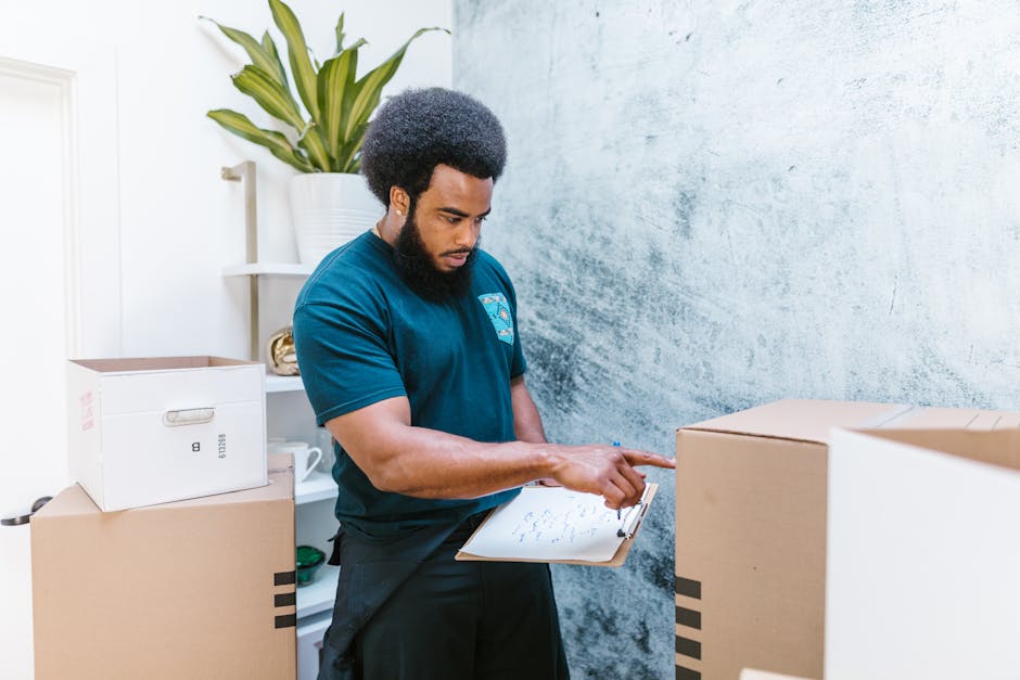 A professional mover from London Storage is inside a room, holding a clipboard and examining packing details. The setting includes several packed cardboard boxes, some sealed with packing tape and oth