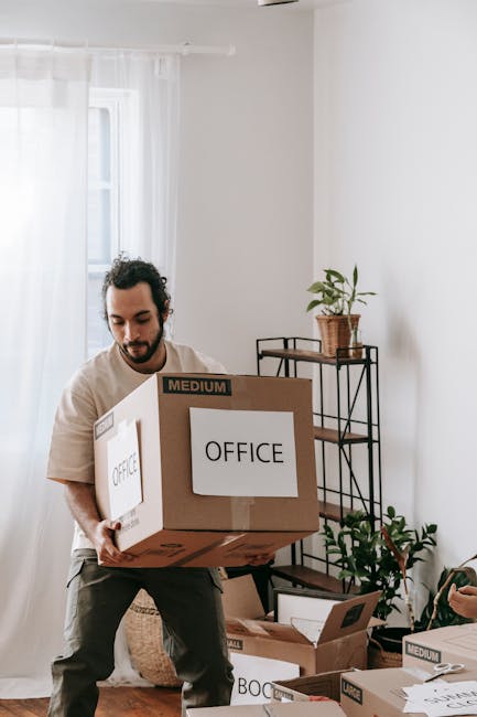 A professional mover from London Storage is inside a room, holding a clipboard and examining packing details. The setting includes several packed cardboard boxes, some sealed with packing tape and oth